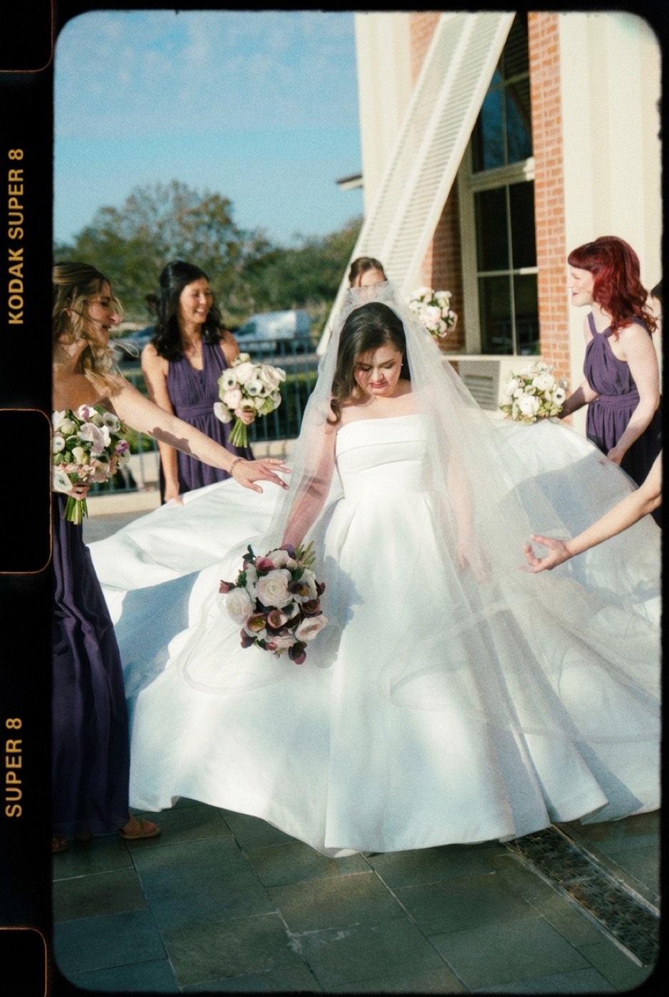 A bride twirls as her bridesmaids fan out her veil and train in afternoon light, framed by a Kodak Super 8 film edge, with heavy warm grain and faded color.