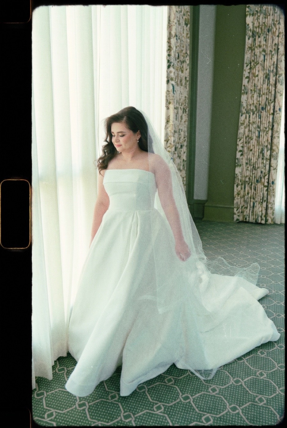 A bride in a long veil stands in soft window light against floral drapes, rendered in warm, grainy, faded Super 8 tones.